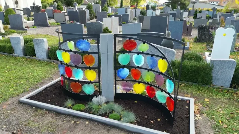 Vorschaubild: zwei geschwungene Stahlflügel mit bunten Glasscheiben und eine helle Stele auf dem Friedhof Lenzfried, von nah gefilmt – STOI Steinmetzwerkstatt