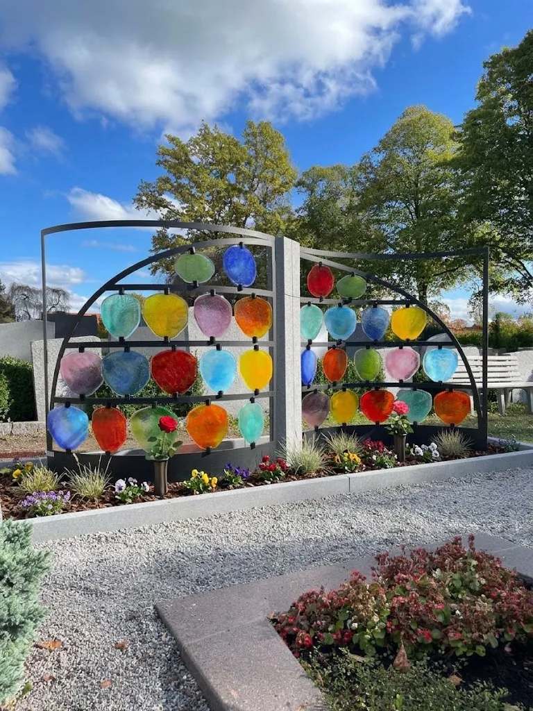 Urnengrabanlage mit bunten Glasscheiben in zwei Bögen, davor Blumen und Kies in der Sonne – STOI Steinmetzwerkstatt