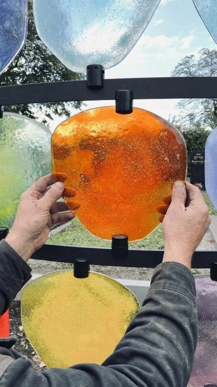 Hände nehmen eine leere orange Glasscheibe aus dem Rahmen der Urnengrabanlage – STOI Steinmetzwerkstatt