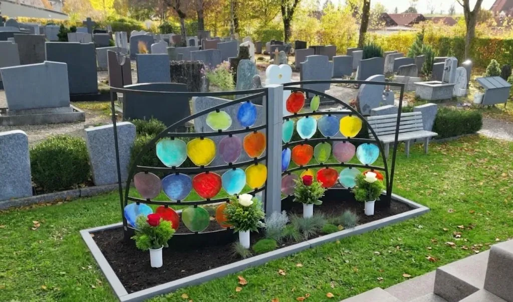Urnengrabfeld HIMMELREICH MEMORIAL von vorne mit bunten Glasscheiben in zwei Bögen, davor Blumen, zwischen Grabsteinen auf dem Friedhof Lenzfried – STOI Steinmetzwerkstatt