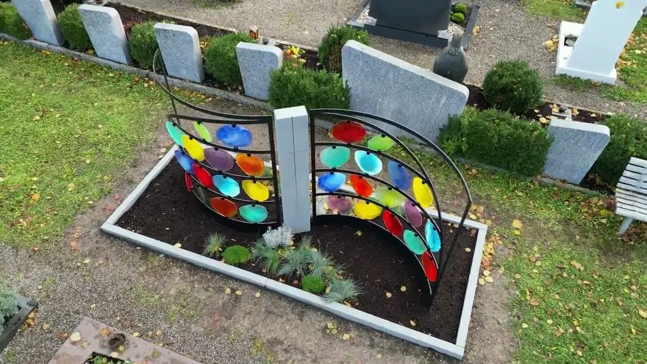 Zwei geschwungene Stahlflügel mit bunten Glasscheiben und eine helle Stele zwischen Grabsteinen auf dem Kemptener Friedhof Lenzfried – STOI Steinmetzwerkstatt